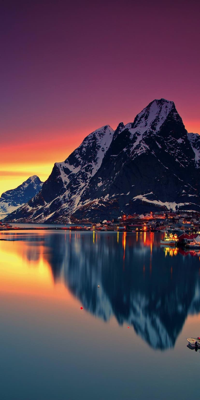 Aurora borealis in pink and orange hues above a snowy mountain and glowing village reflected in a still fjord.