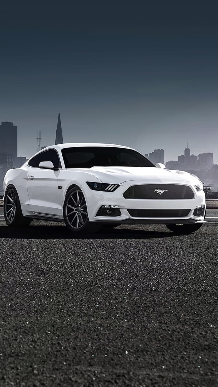 White Ford Mustang GT with a prominent pony emblem parked on asphalt with a blurred city skyline background.