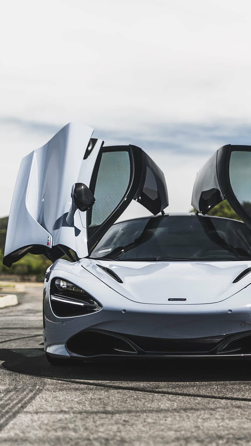 McLaren 720S supercar with silver paint and open dihedral doors against a background of sky and green trees.