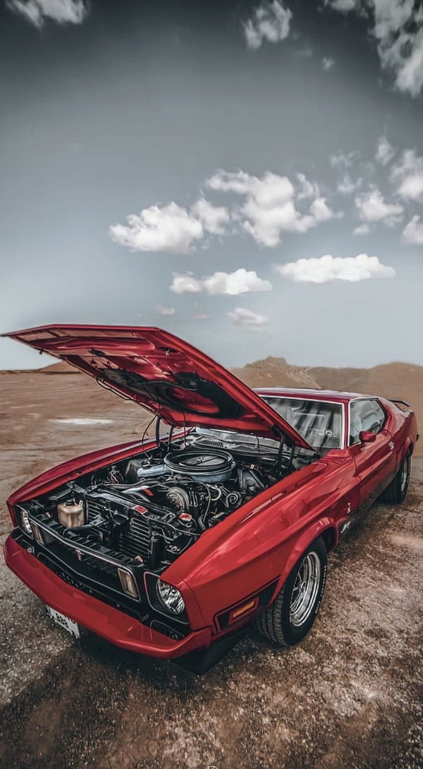 Red Ford Mustang with an open hood reveals its engine in a vast desert landscape under an overcast sky.