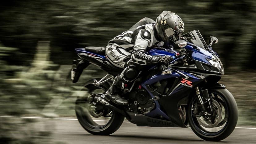 Blue Suzuki GSX-R sportbike leaning into a sharp turn with a motion-blurred forest background on a paved road.