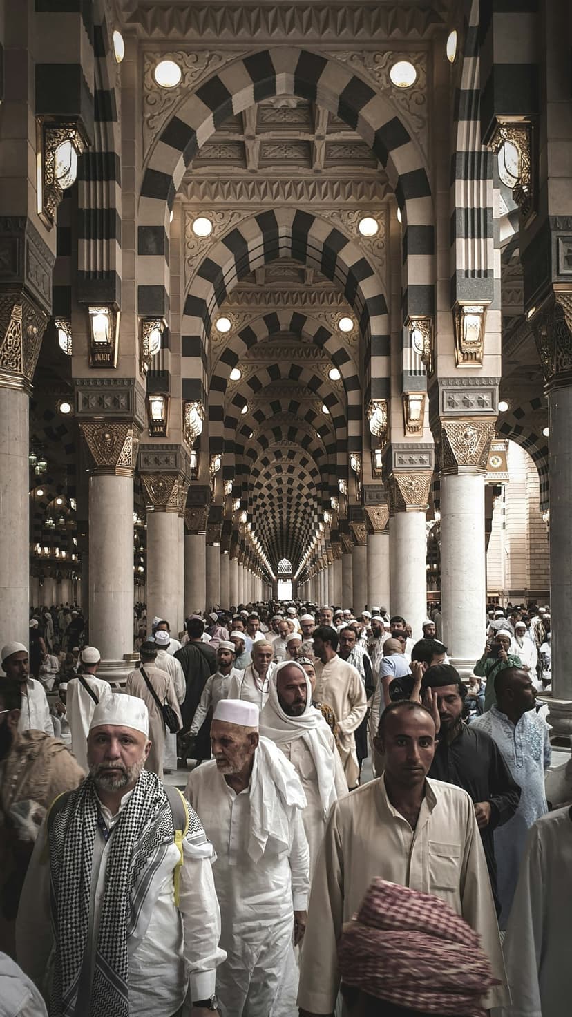 Muslim pilgrims in white ihram walk through the black and white striped arches of the Prophet's Mosque in Medina.