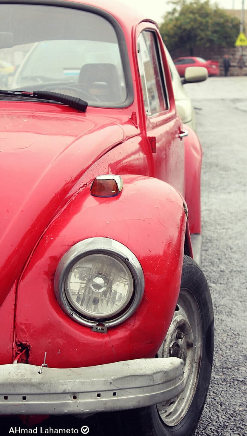 Red vintage Volkswagen Beetle headlight and weathered chrome bumper reflecting on wet asphalt after rain.