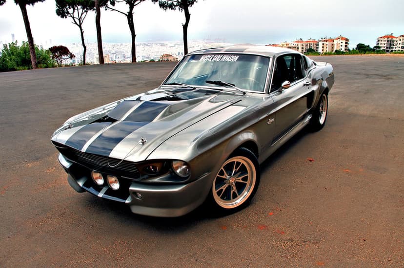 Silver Ford Mustang Shelby GT500 Eleanor with black racing stripes parked on asphalt overlooking a city.
