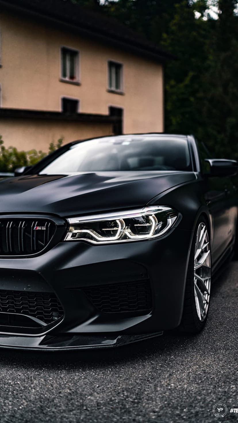 BMW M5 matte black front grille and glowing LED headlights on a grainy asphalt road from a low-angle view.