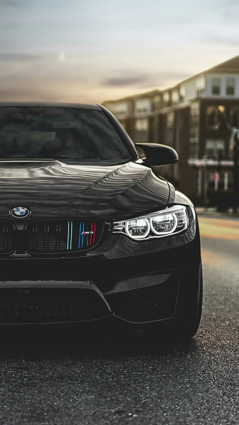 Black BMW M4 coupe with tricolor grille stripes and bright headlights parked in a blurred urban twilight scene.