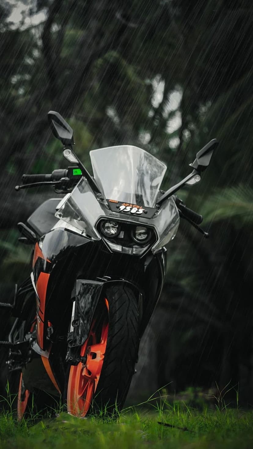 KTM RC motorcycle stands on damp grass under rainfall with orange accents and visible water droplets on the body.