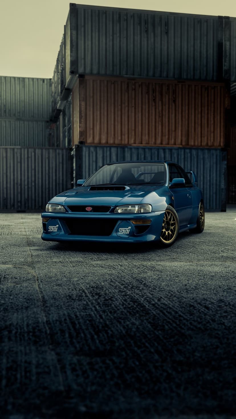 Blue Subaru Impreza 22B STI with gold wheels parked in front of stacked shipping containers on asphalt.