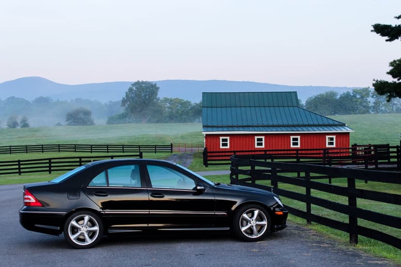 Black Mercedes-Benz sedan on a driveway next to a red barn and misty mountains during a foggy rural sunrise.
