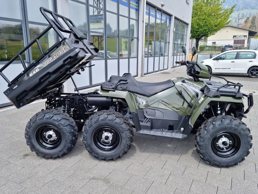 Olive green Polaris 6x6 Big Boss ATV with a raised black cargo bed parked on a paved outdoor area.