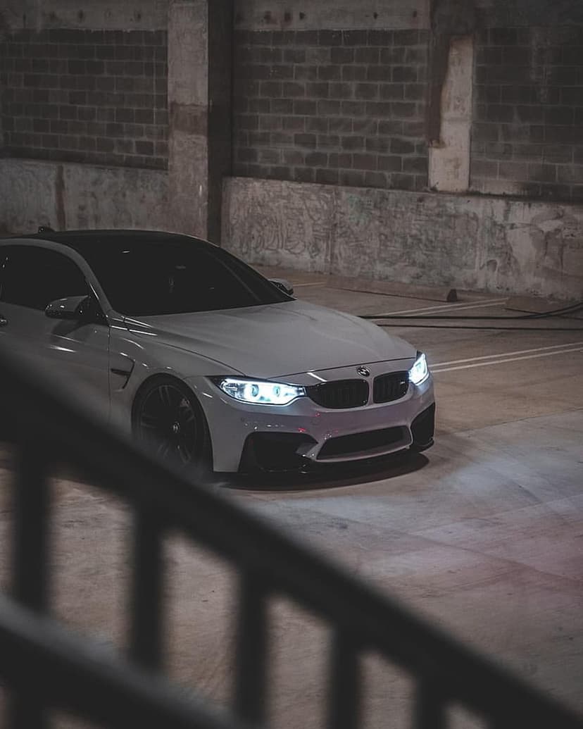 White BMW M4 sports coupe with glowing LED headlights parked against a dark brick wall in an urban garage.