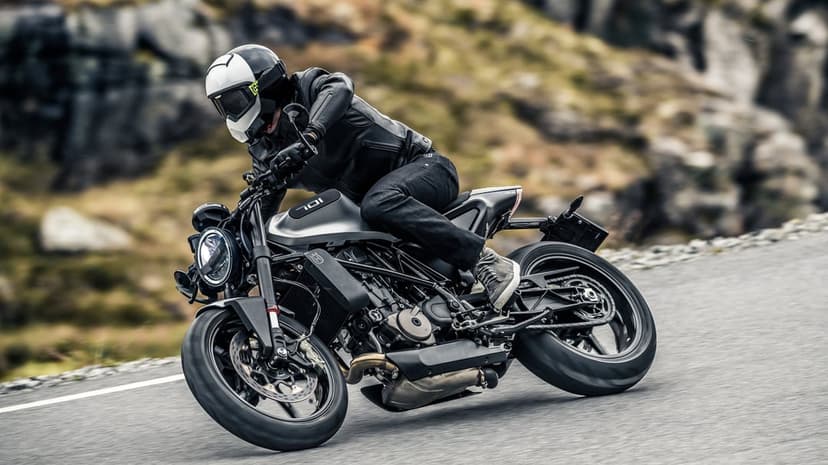 Motorcyclist in black leather leaning a Husqvarna 701 into a sharp turn on a gray asphalt mountain road.