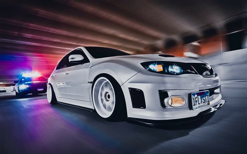 White Subaru WRX STI hatchback speeds through a tunnel with a police car in pursuit under flashing emergency lights.