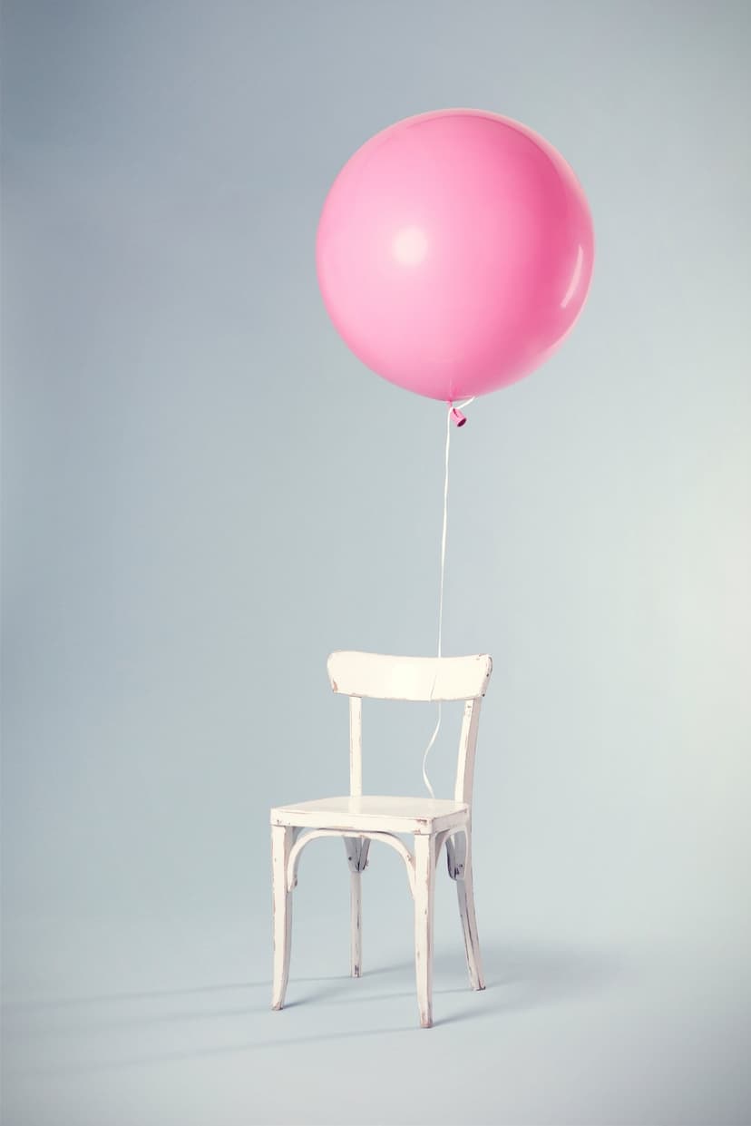 Pink balloon floats tethered to a rustic white chair against a soft blue-gray minimalist studio backdrop.
