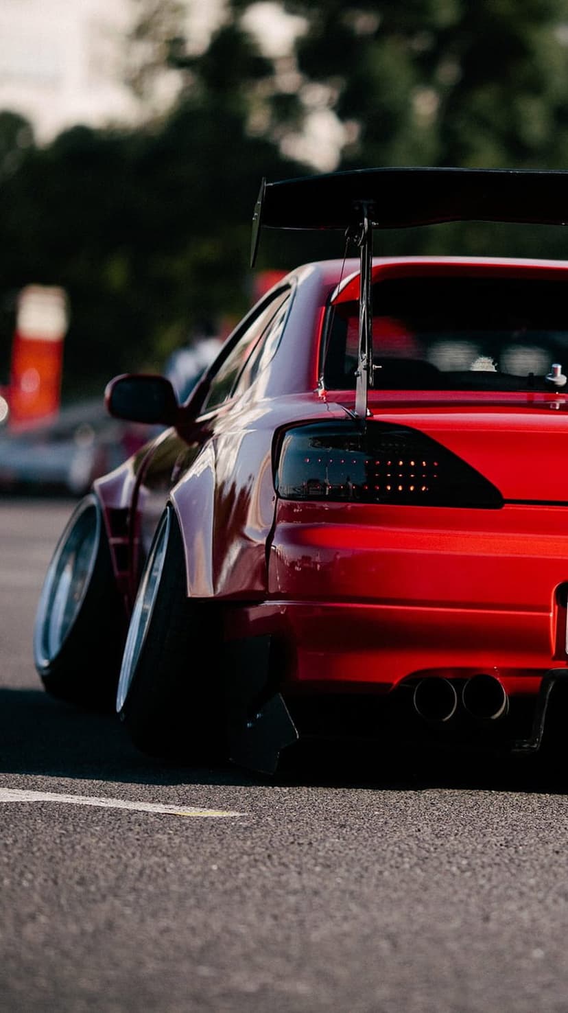 Red sports car rear view showing a large black spoiler, widebody fender flares, and dual exhaust pipes.