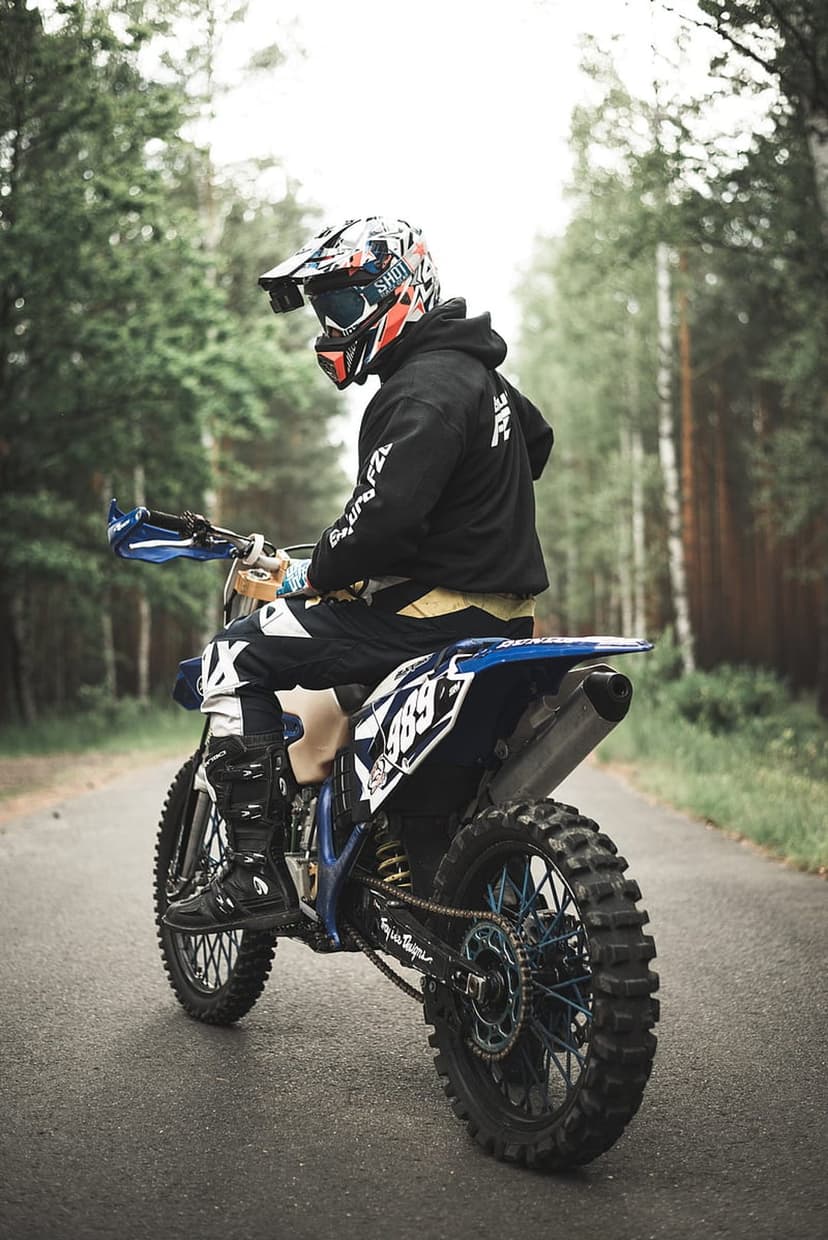 Motocross rider in full gear sits on a blue dirt bike on a forest road with tall pine trees in the background.