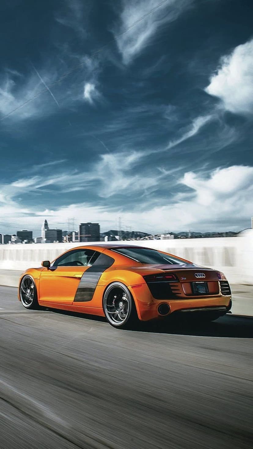 Orange Audi R8 supercar speeds on a highway with a blurred city skyline and cloudy sky in the background.