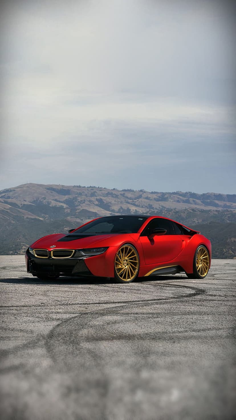 Red BMW i8 with gold rims on a textured grey road featuring tire marks with rolling hills in the background.