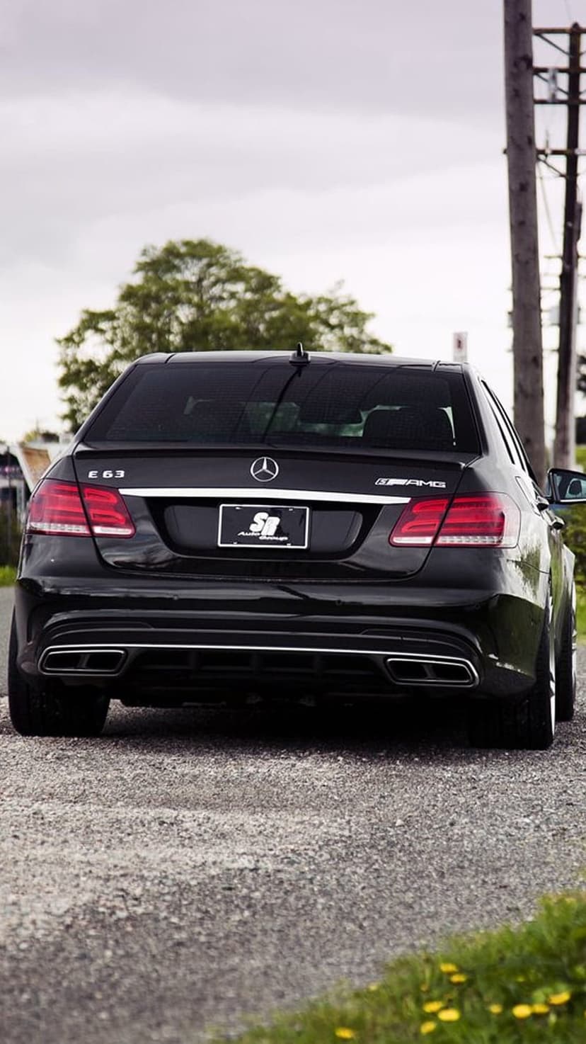 Mercedes-Benz E63 AMG rear view with quad exhausts on a gravel road under a cloudy sky during the daytime.