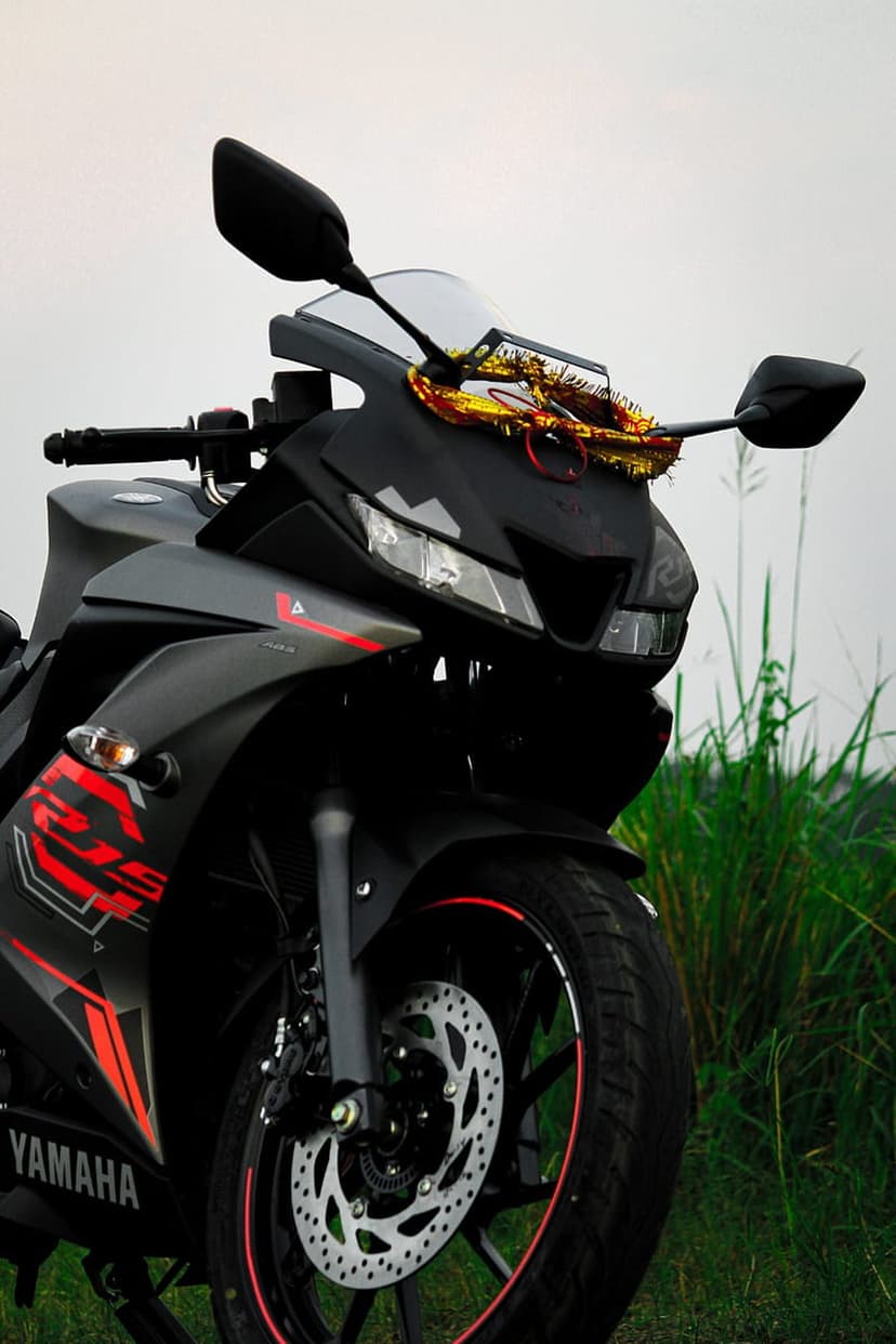 Yamaha R15 black sportbike with a floral garland draped over the fairing, set against green grass and blue sky.