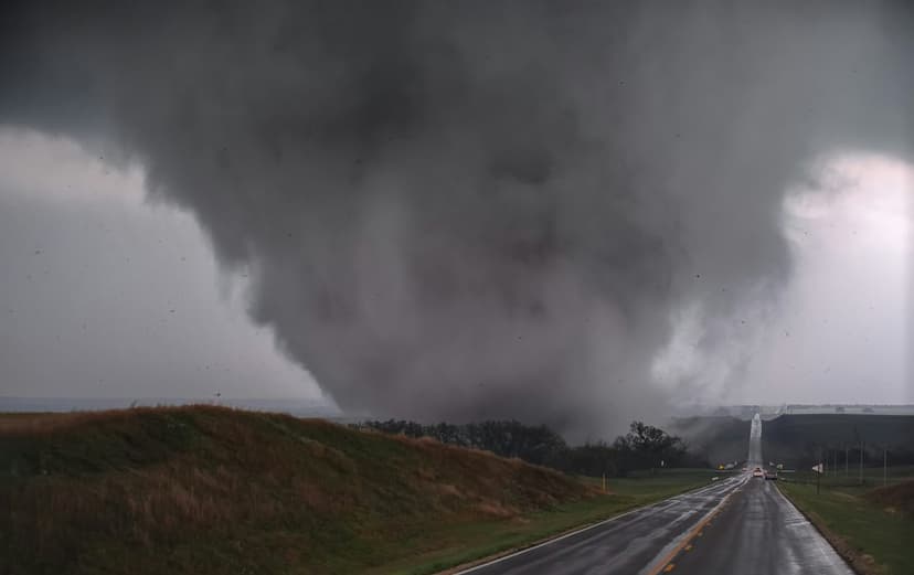 Large dark funnel cloud descends onto a wet highway surrounded by green fields and trees under storm clouds.