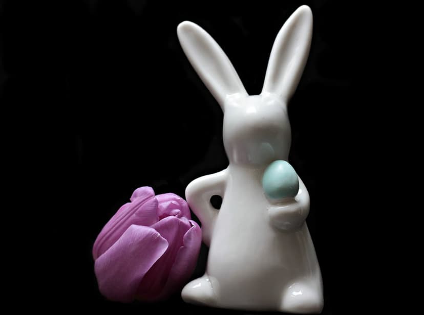 White ceramic rabbit figurine holds a blue egg next to a purple tulip against a dark, minimalist background.