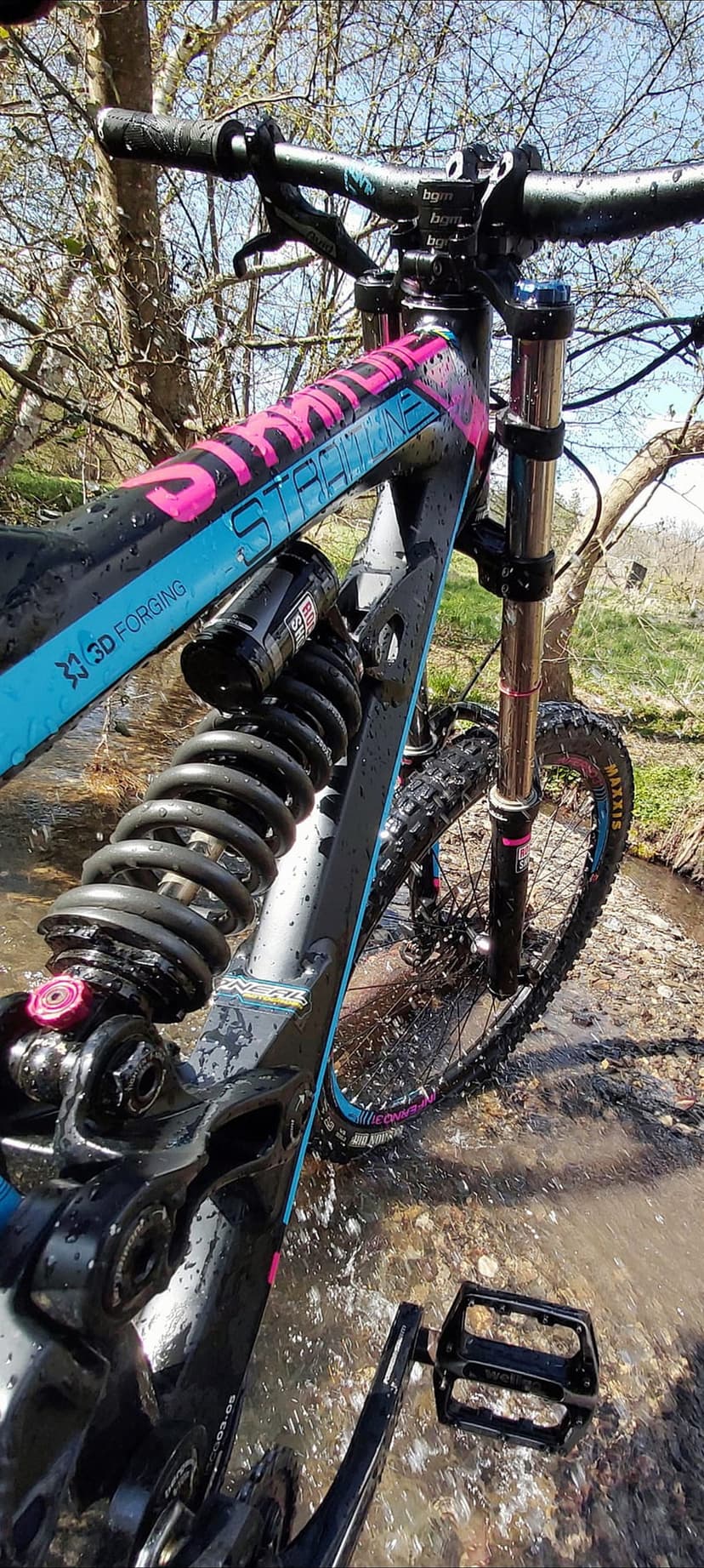 Downhill mountain bike frame with pink and blue graphics sits in shallow splashing water with visible coil shock.