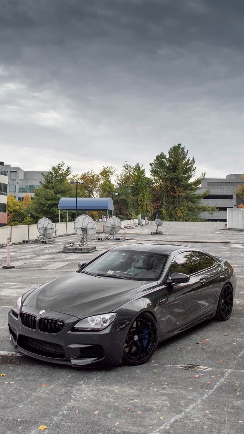 BMW M6 coupe in dark grey with black rims and blue calipers parked on a rooftop lot under a cloudy sky.