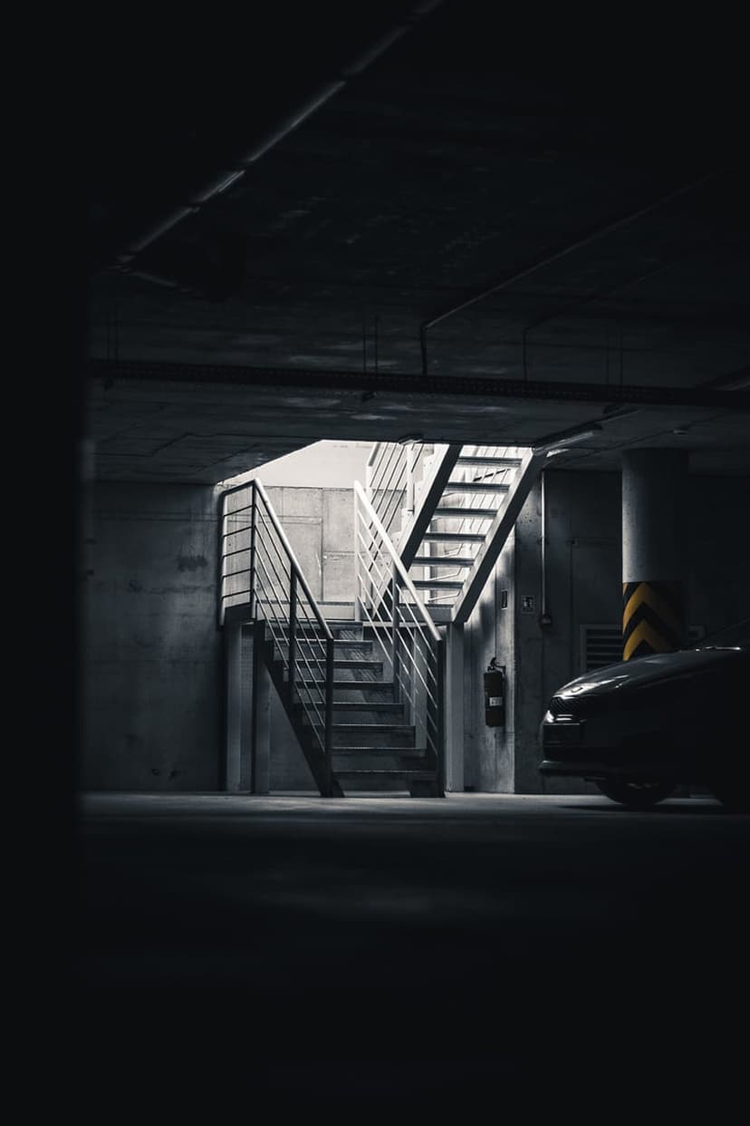 Metal stairwell in a dark concrete parking garage with a car partially visible under harsh overhead lighting.