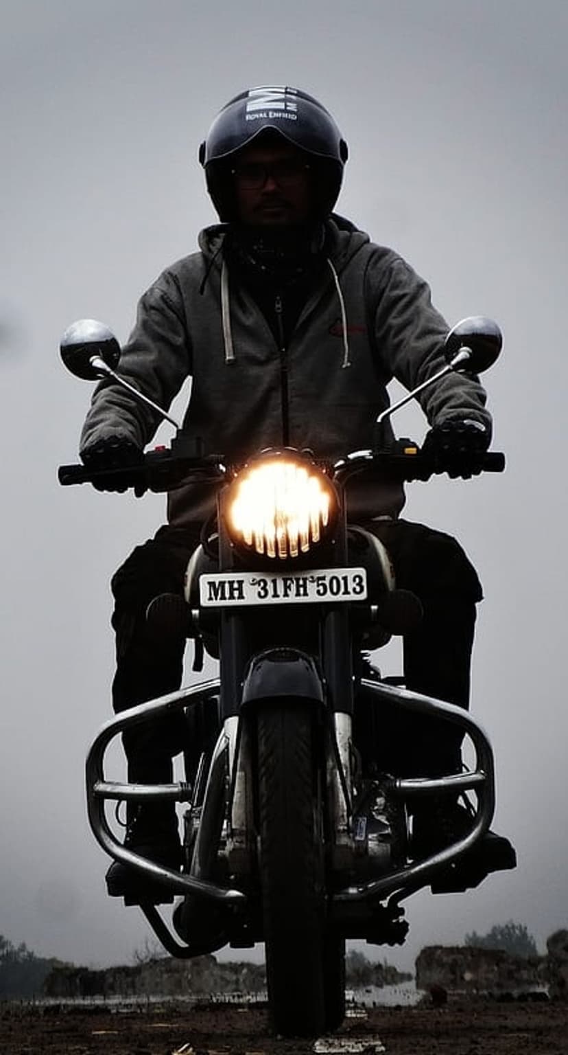 Biker in a black helmet and grey hoodie rides a Royal Enfield motorcycle with its headlight on over rocky soil.