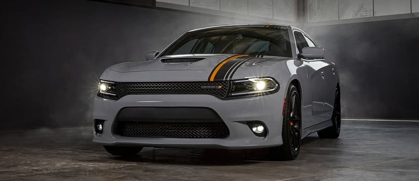 Grey Dodge Charger muscle car with orange stripes and glowing headlights in a dark, smoky studio environment.