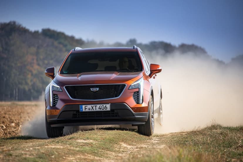 Front view of an orange Cadillac XT4 driving on a dirt path, kicking up a large cloud of dust under a blue sky.