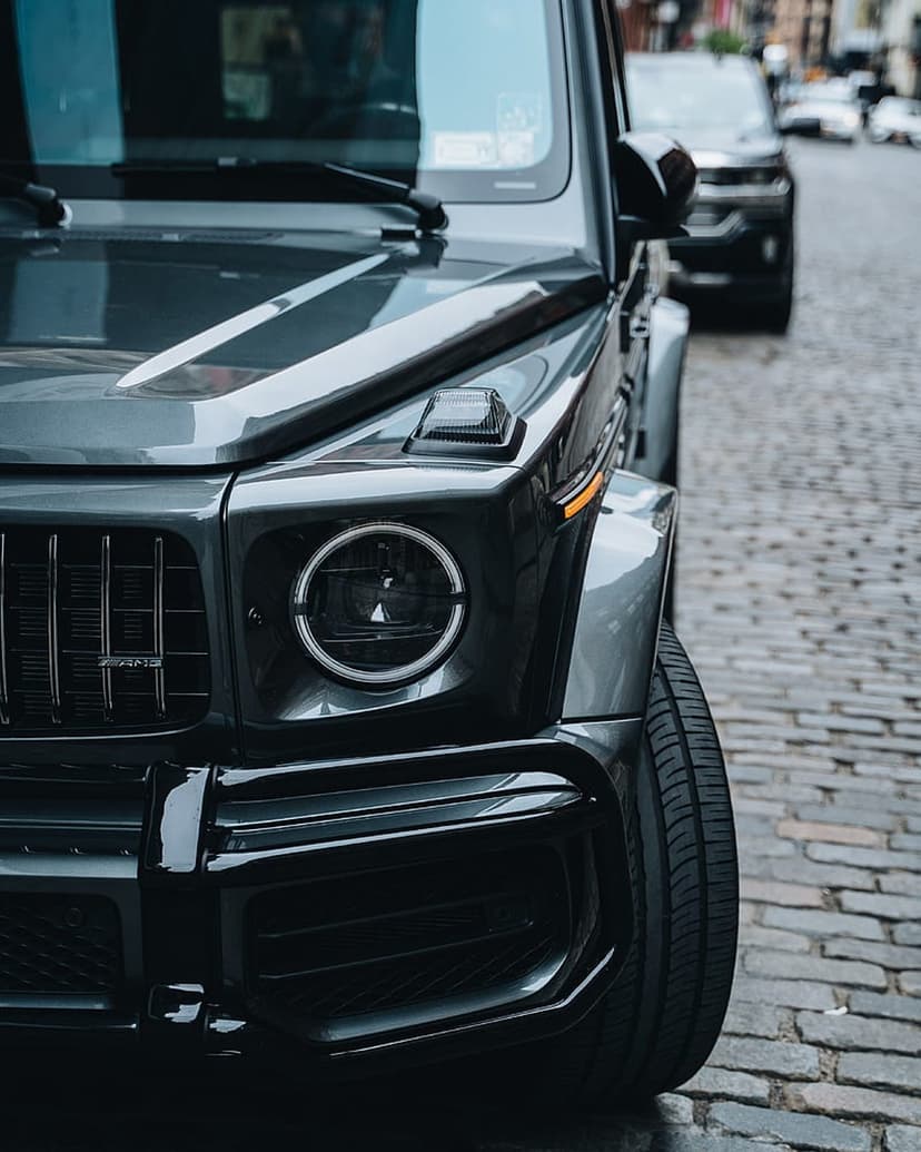 Mercedes-AMG G 63 front profile with round LED headlights and dark gray paint on a textured cobblestone street.