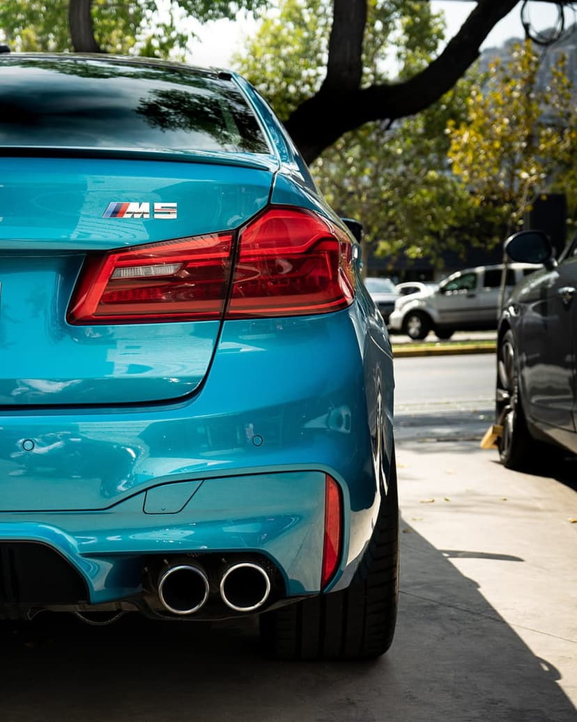 Aqua BMW M5 rear bumper featuring quad exhausts, M5 trunk badge, and glowing red LED taillights on a paved road.