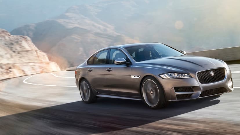 Grey Jaguar sedan driving on a winding mountain road with motion blur and sun-drenched peaks in the background.