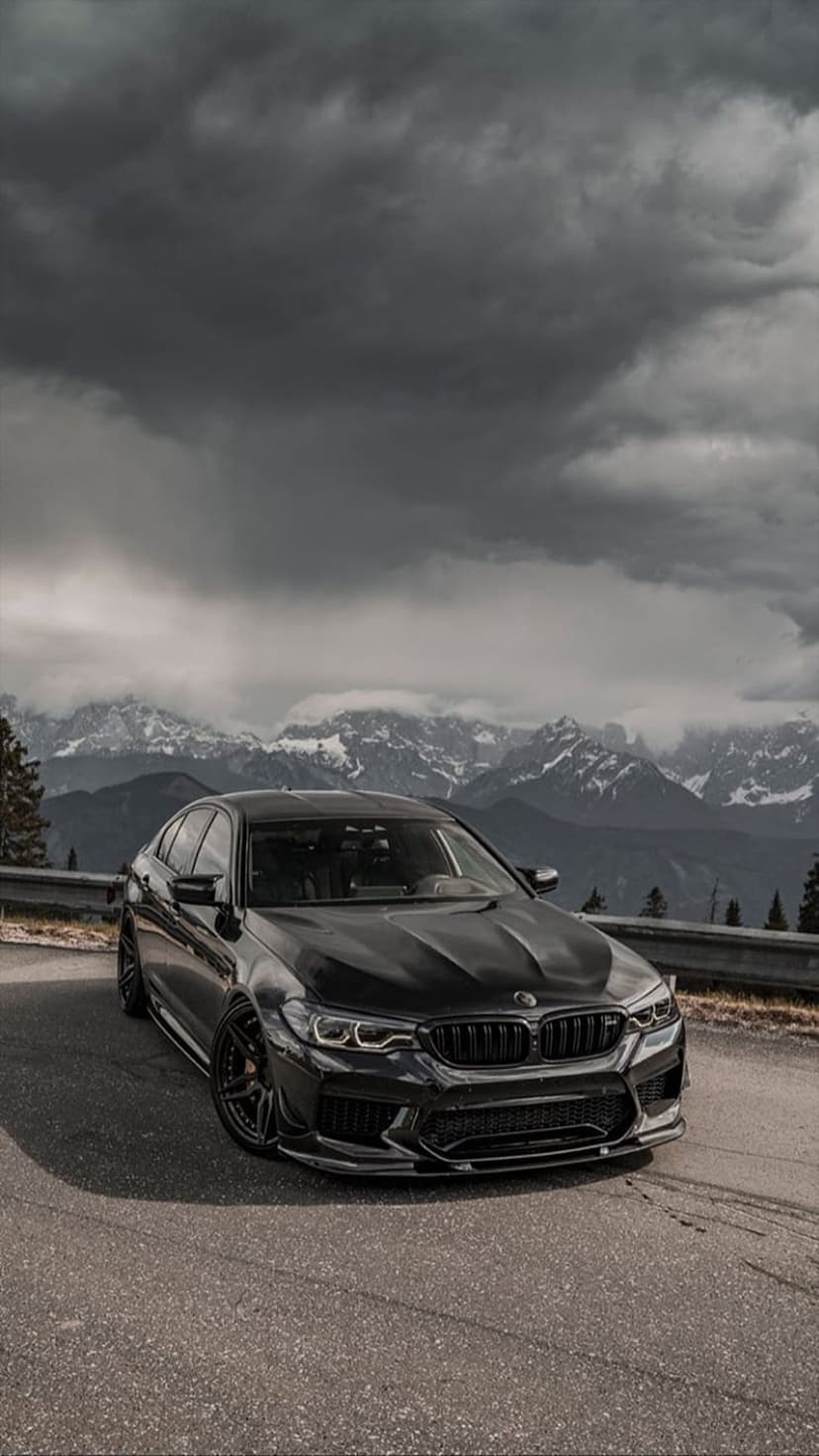 Black BMW M5 sedan parked on a mountain road with snow-capped peaks beneath dark, dramatic storm clouds.