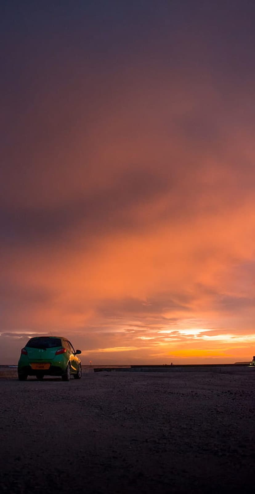 Green hatchback car parked on a dark pebble beach against a vibrant orange and purple sunset over the ocean.