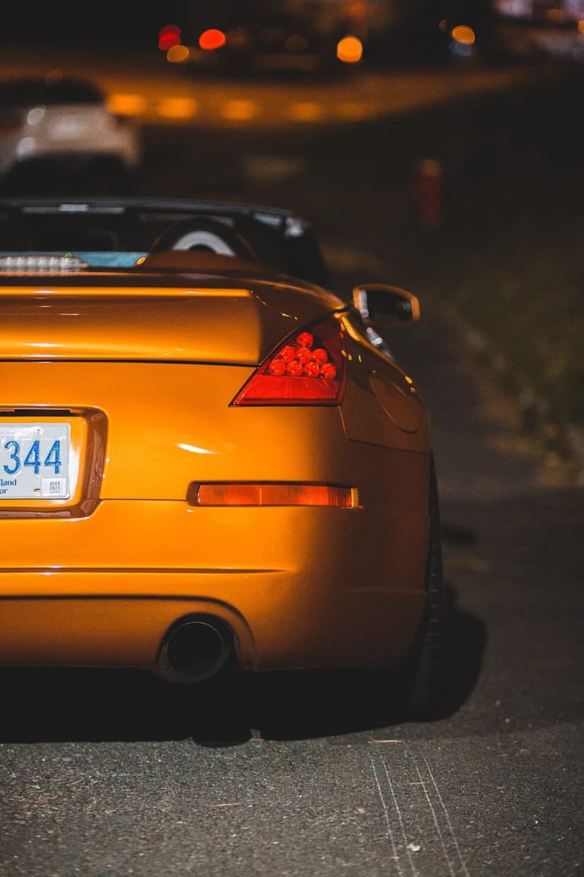 Orange convertible car rear with lit red taillights and exhaust pipes against blurred night city lights.