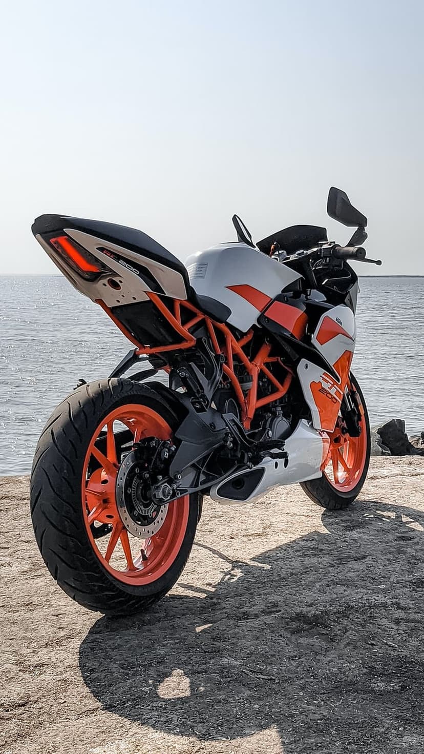 White and orange KTM RC sportbike parked on jagged shore rocks overlooking a calm blue sea under a clear sky.