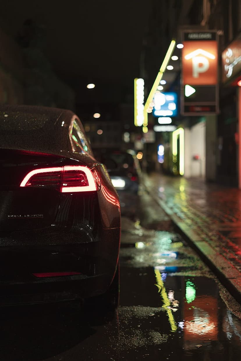 Red Tesla taillights glowing on a wet city street at night with neon reflections on the damp pavement.