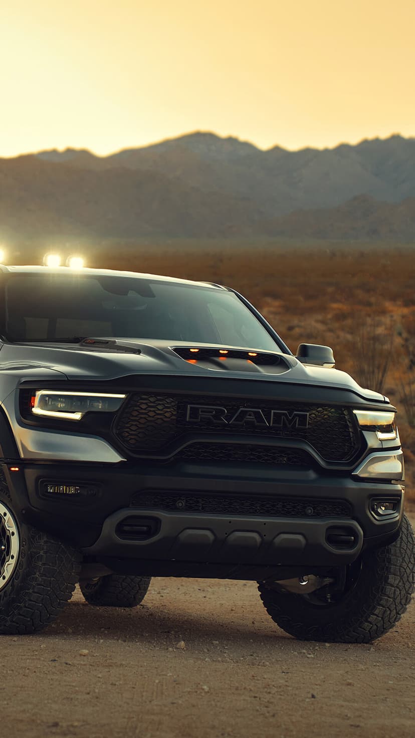 Front view of a dark grey RAM TRX truck in a desert with mountains at sunset and glowing headlights.