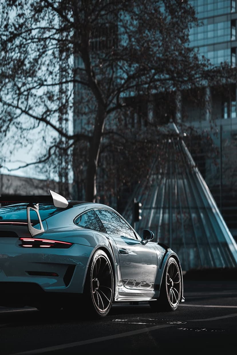 Light blue Porsche 911 GT3 RS with a rear wing parked near a glass building and a tree on a dark urban street.