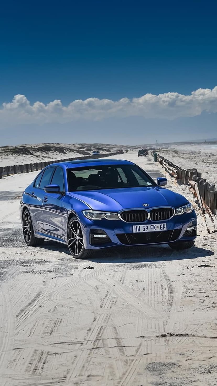 Blue BMW 3 Series sedan driving on a sandy beach road between wooden fences under a clear blue summer sky.