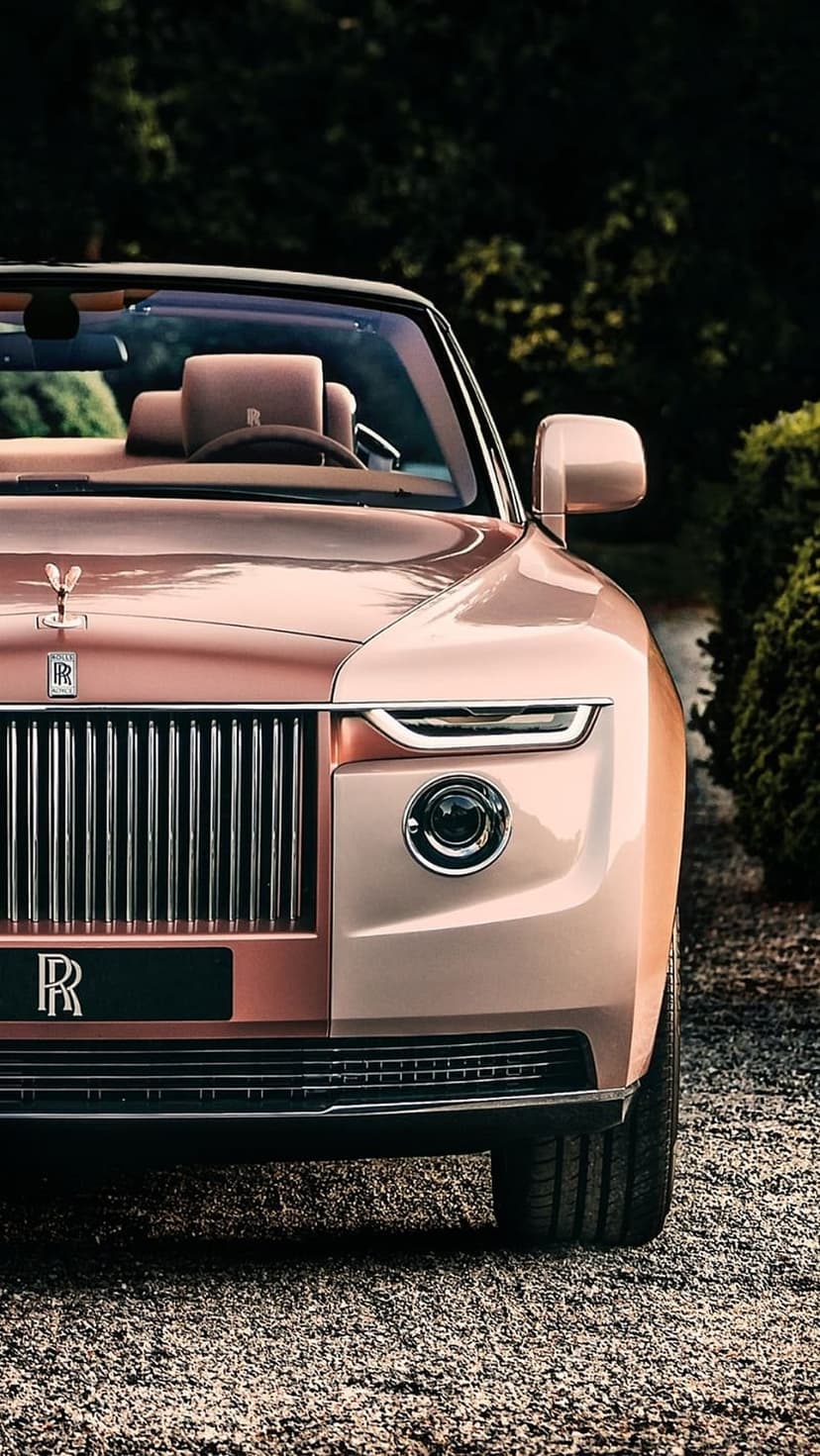 Rose gold Rolls-Royce convertible with a chrome grille and Spirit of Ecstasy ornament on a gravel driveway.
