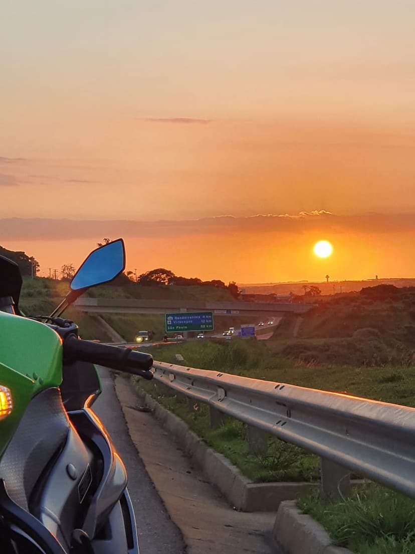 Motorcycle rider standing by a highway overlooking a bright orange sunset and lush green forest landscape.