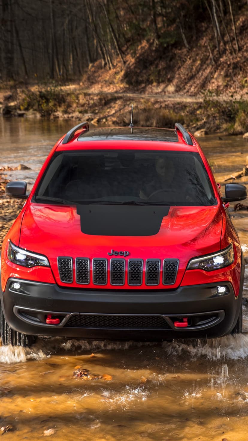 Red Jeep Cherokee Trailhawk driving through a shallow rippling stream in a forest with autumn foliage.