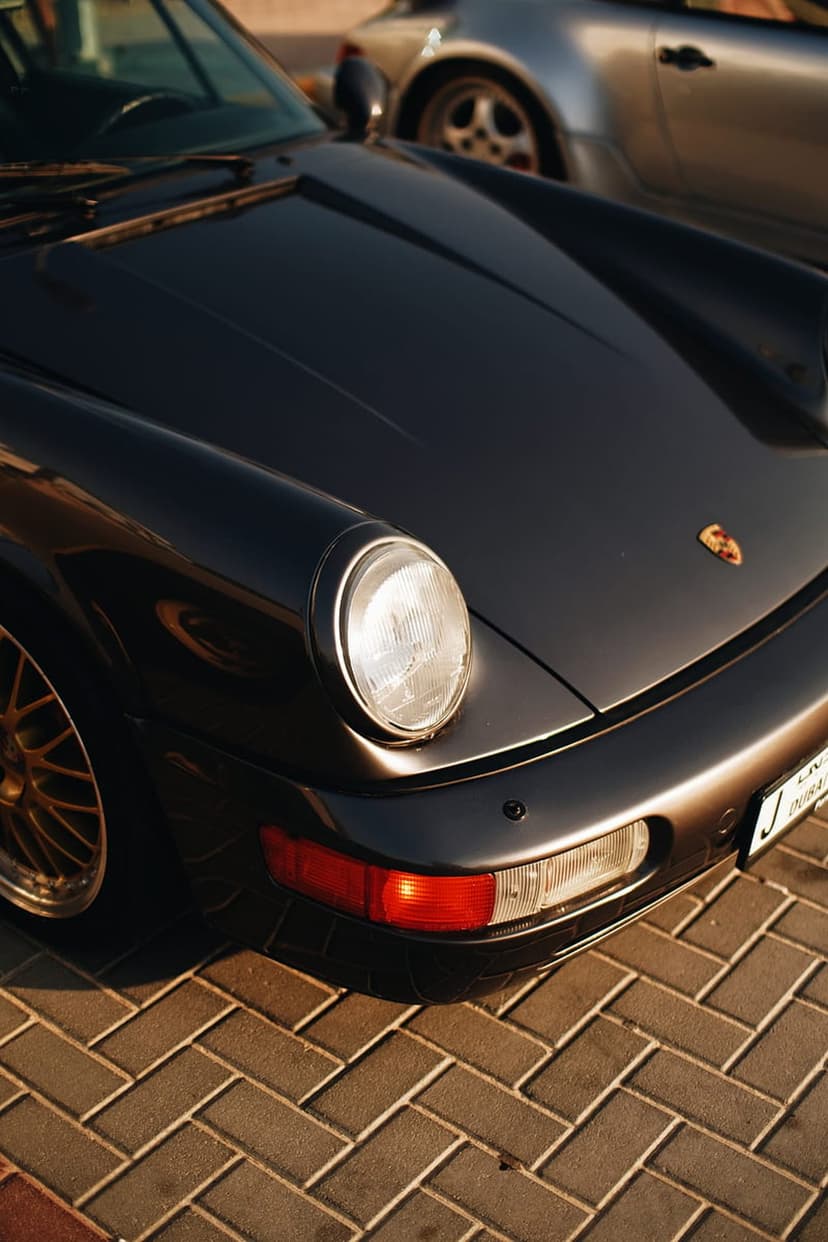 Porsche 911 front fender and headlight featuring a gold hood crest on dark paint over a brick-paved street.
