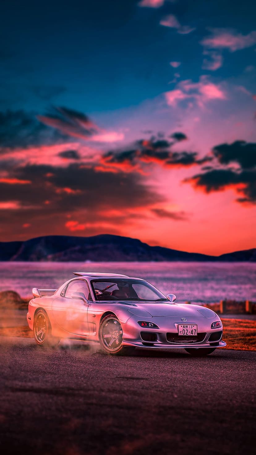 Silver Mazda RX-7 drifting with tire smoke on a coastal road under a vibrant purple and orange sunset.