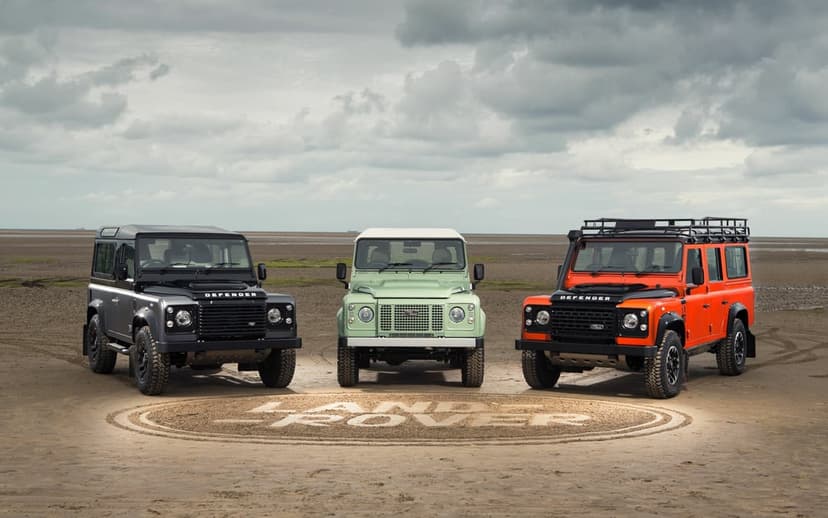 Three Land Rover Defenders in black, mint green, and orange parked on a beach with a logo carved in the sand.
