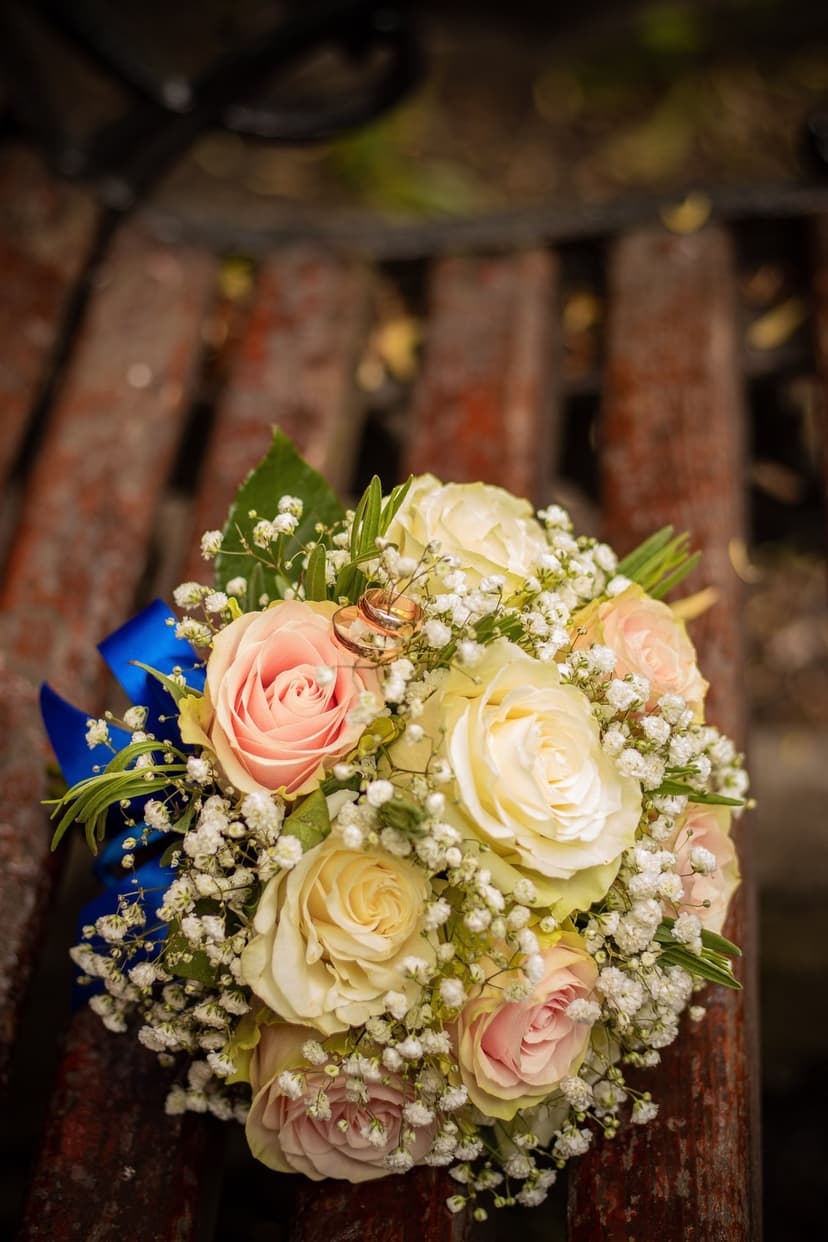 Pink roses and gold wedding rings on a rustic wooden bench with a blue ribbon tied around the flower stems.